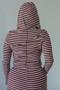 Woman wearing a burgundy and mauve striped patterned set featuring a hoodie top and high waisted pants. The photo is taken indoors against a white wall. The photo shows the back of the hoodie with the hood up. 