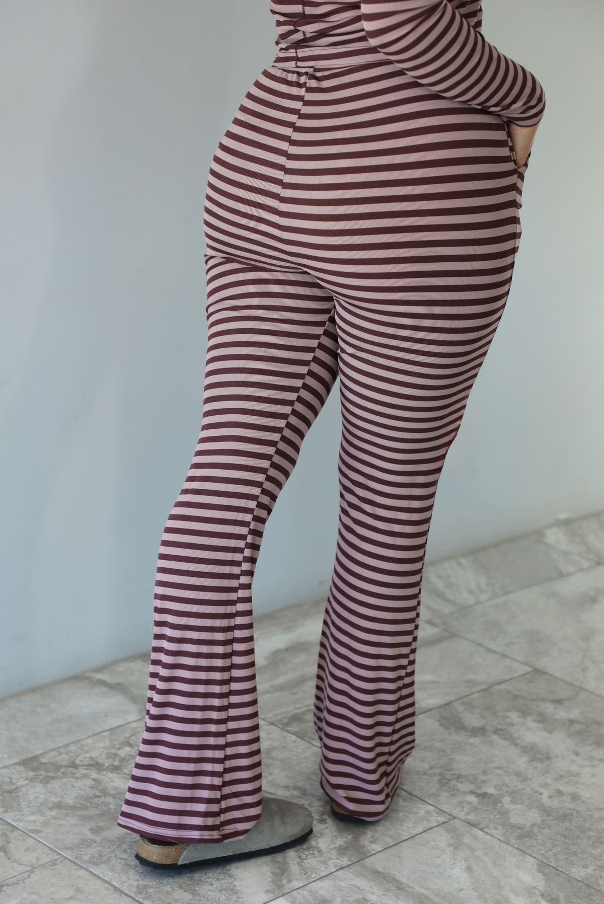 Woman wearing a burgundy and mauve striped patterned set featuring a hoodie top and high waisted pants. The photo is taken indoors against a white wall with white marble flooring. The photo shows a closeup of the back of the pants. 