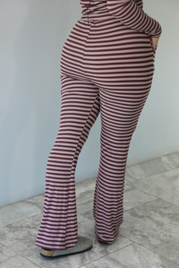 Woman wearing a burgundy and mauve striped patterned set featuring a hoodie top and high waisted pants. The photo is taken indoors against a white wall with white marble flooring. The photo shows a closeup of the back of the pants. 