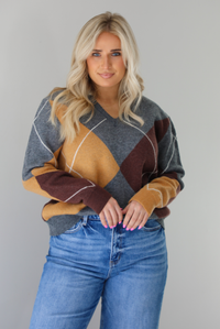 Woman wearing a patterned sweater and blue jeans. The photo is taken indoors against a white wall. The photo shows a closeup of the sweater, which is front tucked into the denim. 