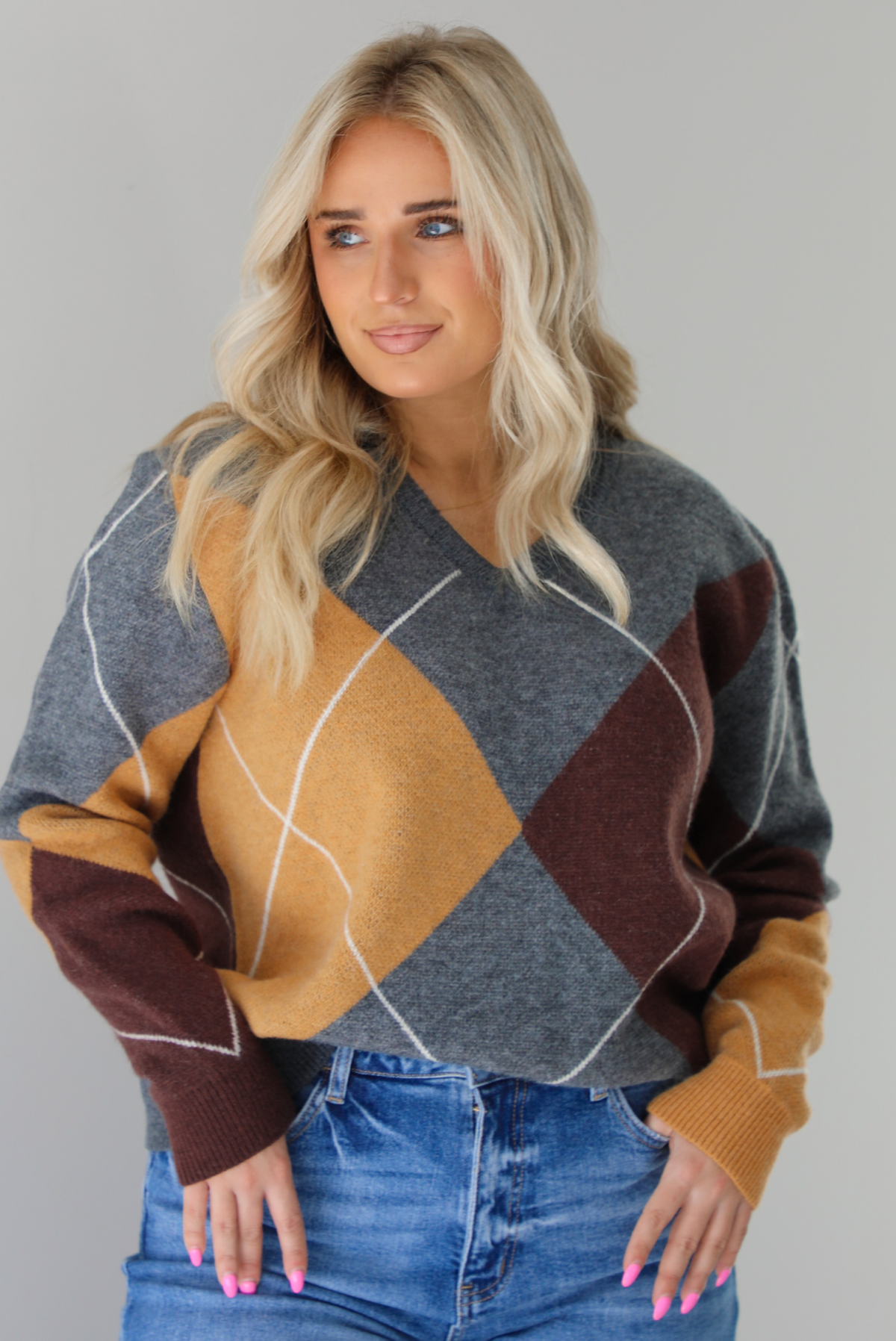 Woman wearing a patterned sweater and blue jeans. The photo is taken indoors against a white wall. The photo shows a closeup of the sweater, which is front tucked into the denim. 