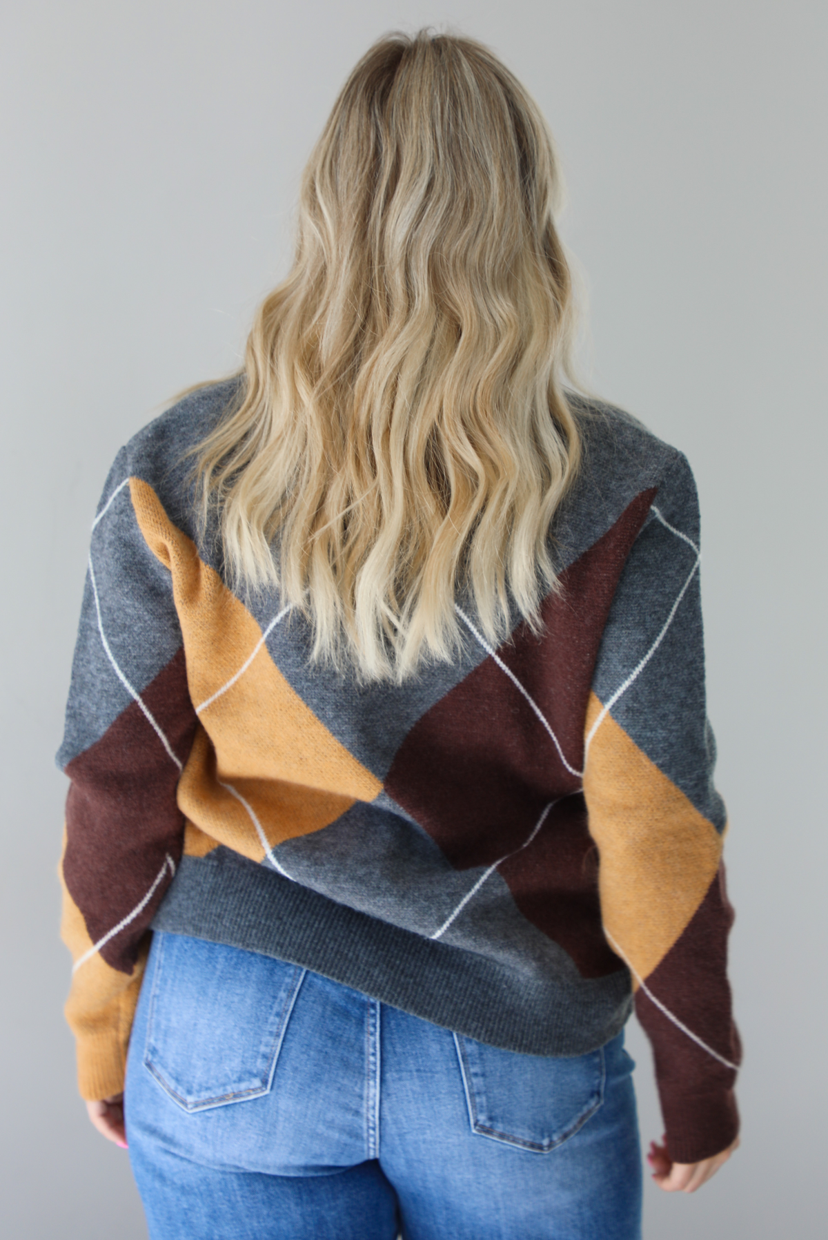 Woman wearing a patterned sweater and blue jeans. The photo is taken indoors against a white wall. The photo shows a closeup of the back of the sweater. 