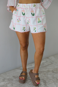 Woman wearing a two piece set that has an embroidered floral print pattern throughout and consists of a collared, oversized short sleeve top and high waisted shorts. The photo is taken indoors against a white wall with white marble flooring and shows the shorts, which are high waisted, have an elastic and drawstring waistline, and have functional pockets on either side. 