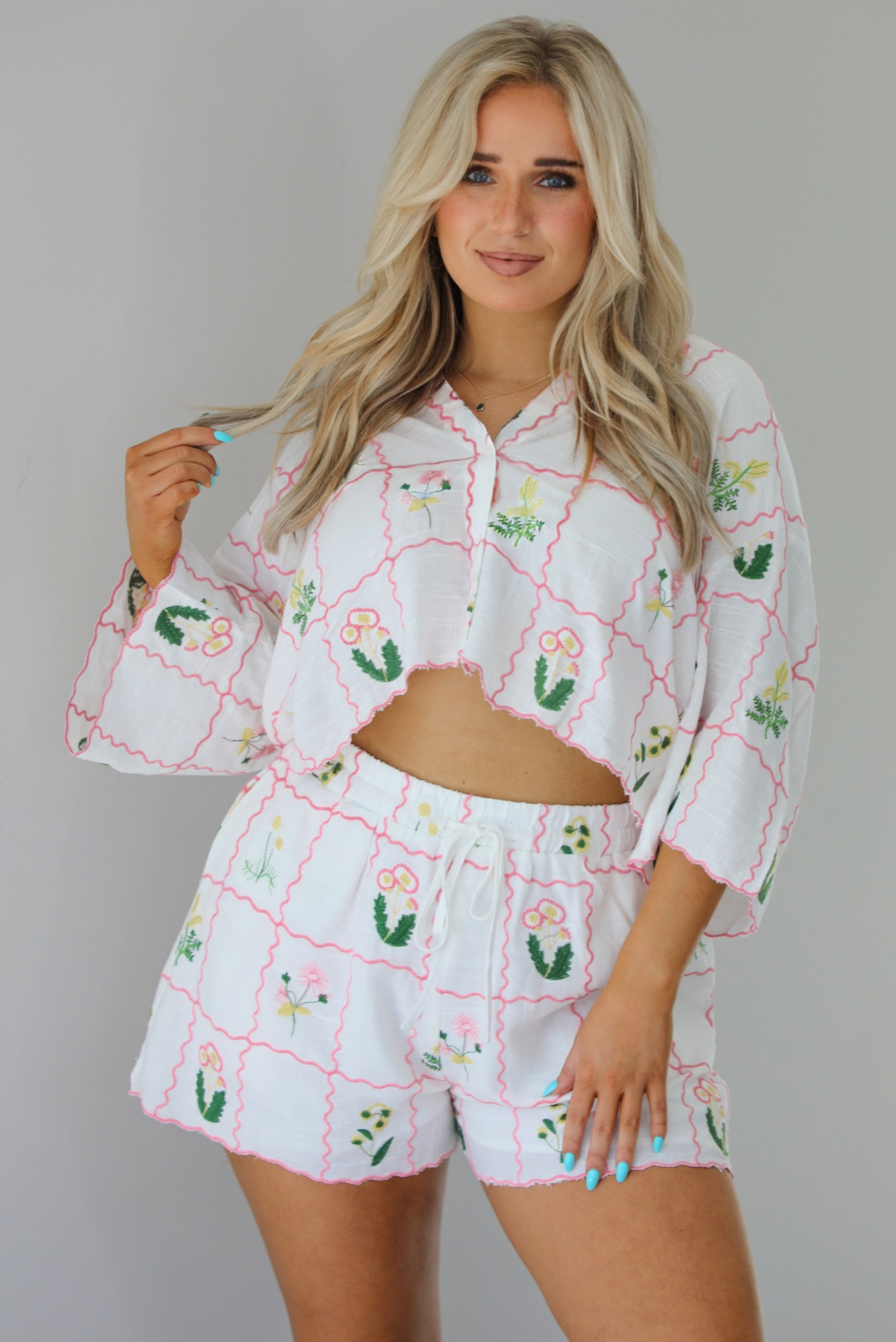Woman wearing a two piece set that has an embroidered floral print pattern throughout and consists of a collared, oversized short sleeve top and high waisted shorts. The photo is taken indoors against a white wall and shows a closeup of the set.
