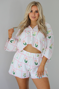 Woman wearing a two piece set that has an embroidered floral print pattern throughout and consists of a collared, oversized short sleeve top and high waisted shorts. The photo is taken indoors against a white wall and shows a closeup of the set.