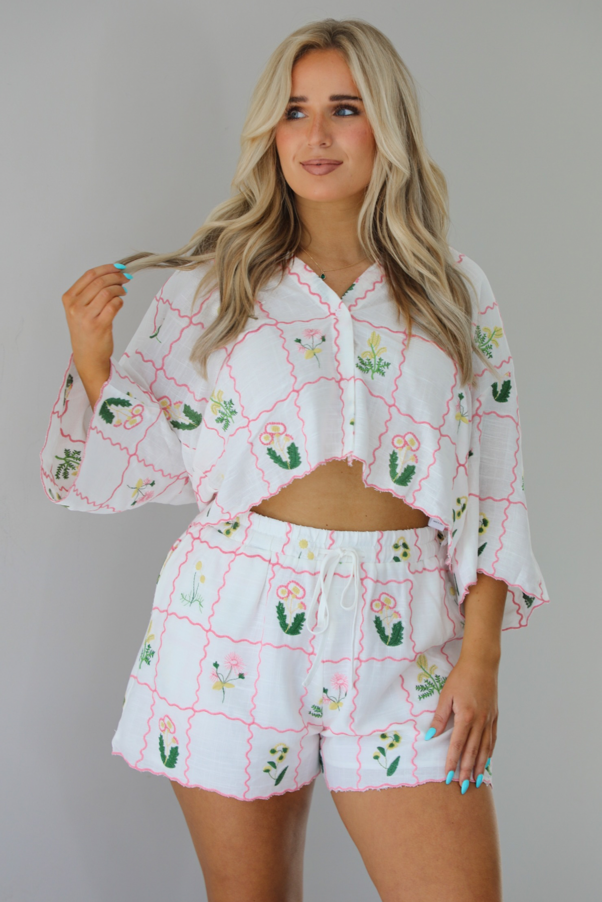 Woman wearing a two piece set that has an embroidered floral print pattern throughout and consists of a collared, oversized short sleeve top and high waisted shorts. The photo is taken indoors against a white wall and shows a closeup of the set.