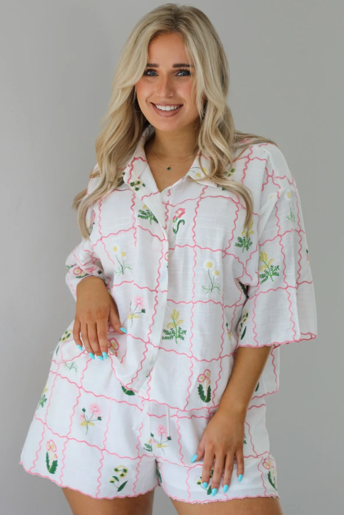 Woman wearing a two piece set that has an embroidered floral print pattern throughout and consists of a collared, oversized short sleeve top and high waisted shorts. The photo is taken indoors against a white wall and shows a closeup of the set.