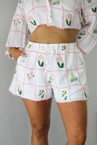 Woman wearing a two piece set that has an embroidered floral print pattern throughout and consists of a collared, oversized short sleeve top and high waisted shorts. The photo is taken indoors against a white wall and shows the shorts, which are high waisted, have an elastic and drawstring waistline, and have functional pockets on either side. 