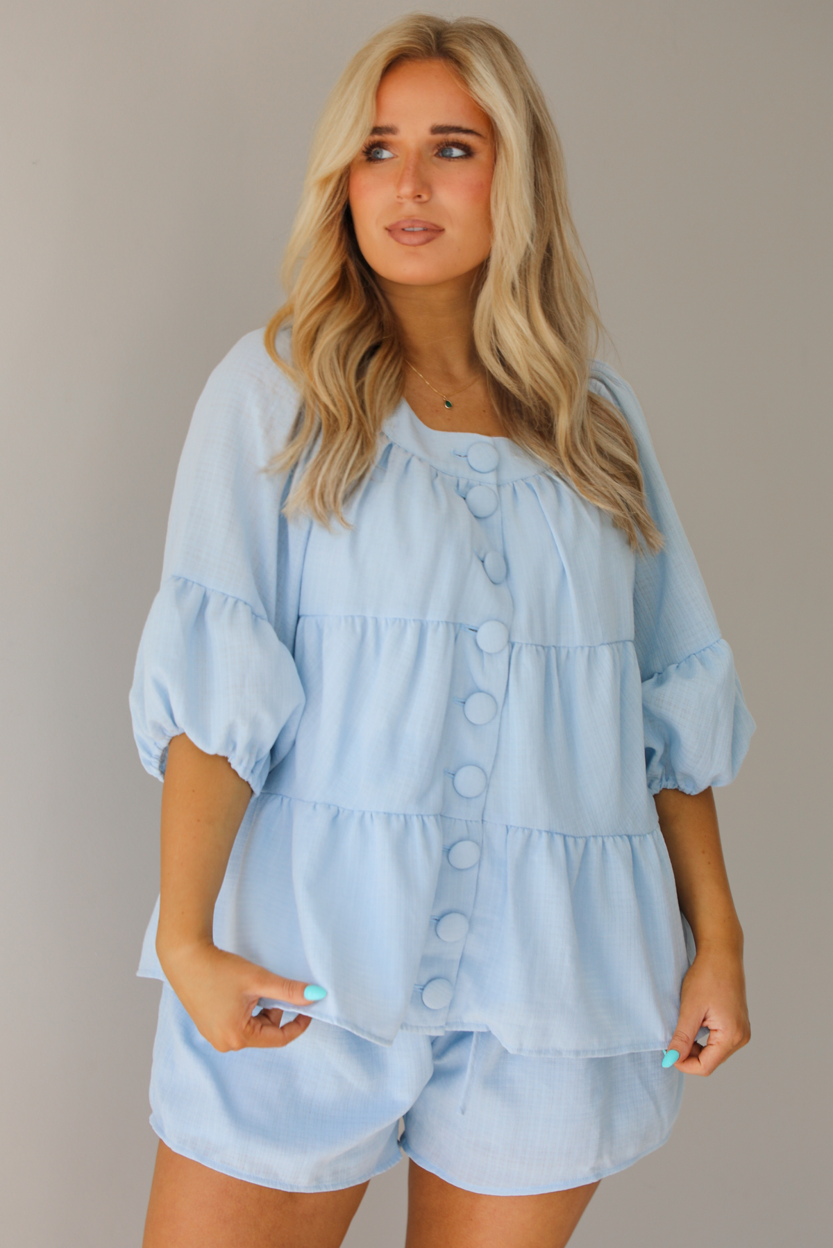 Woman wearing a blue two piece set featuring an oversized top with non-functional button details in the front and shorts. The photo is taken indoors against a white wall, and shows a closeup of the top, which has quarter length sleeves and seam details giving the top a tiered look.