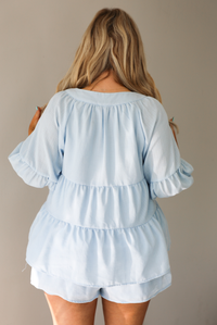 Woman wearing a blue two piece set featuring an oversized top with non-functional button details in the front and shorts. The photo is taken indoors against a white wall, and shows a closeup of the back of the top, which has quarter length sleeves and seam details giving the top a tiered look.