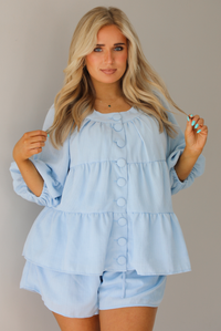 Woman wearing a blue two piece set featuring an oversized top with non-functional button details in the front and shorts. The photo is taken indoors against a white wall, and shows a closeup of the top, which has quarter length sleeves and seam details giving the top a tiered look.