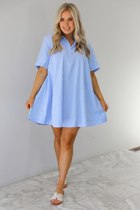 Woman wearing a blue and white striped dress that has a collared neckline, functional quarter button closures in the front, and a flowy skirt. The photo is taken indoors against a white wall with white marble flooring. 