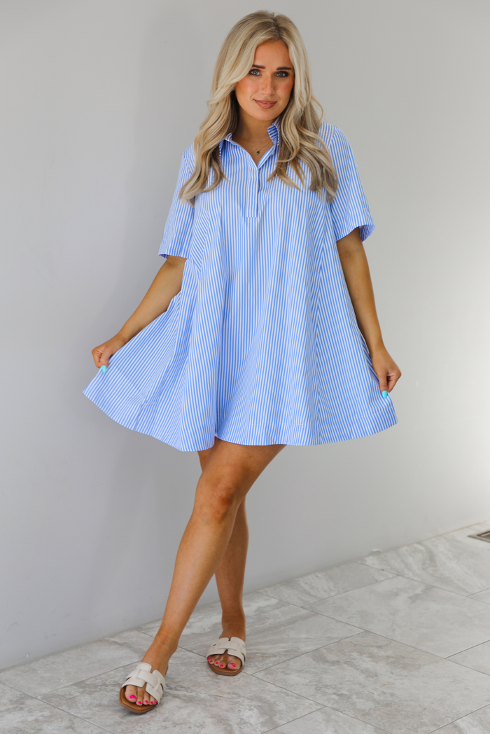 Woman wearing a blue and white striped dress that has a collared neckline, functional quarter button closures in the front, and a flowy skirt. The photo is taken indoors against a white wall with white marble flooring. 
