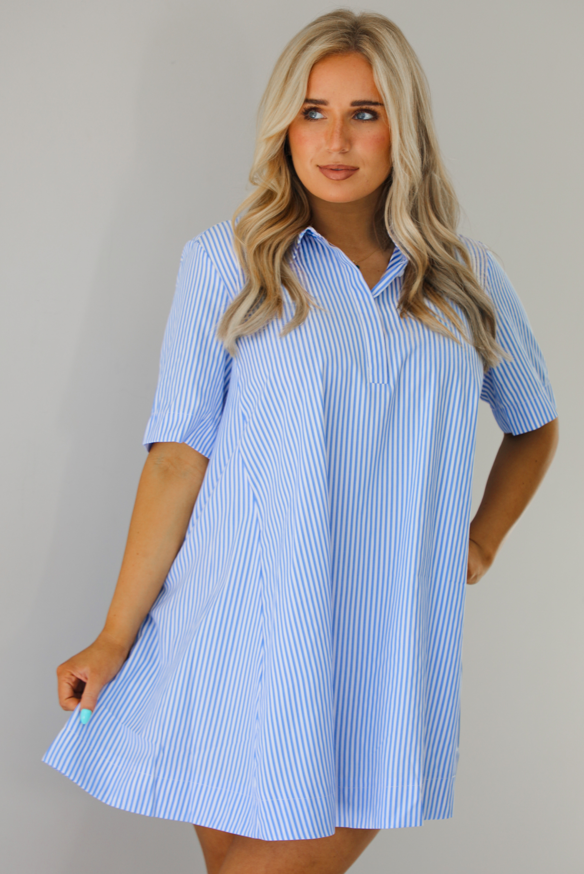 Woman wearing a blue and white striped dress that has a collared neckline, functional quarter button closures in the front, and a flowy skirt. The photo is taken indoors against a white wall.