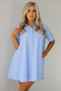 Woman wearing a blue and white striped dress that has a collared neckline, functional quarter button closures in the front, and a flowy skirt. The photo is taken indoors against a white wall.