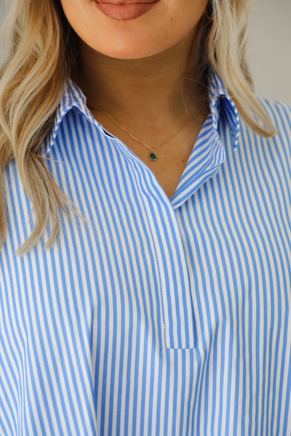 Woman wearing a blue and white striped dress that has a collared neckline, functional quarter button closures in the front, and a flowy skirt. The photo is taken indoors against a white wall and shows a closeup of the collared neckline. 