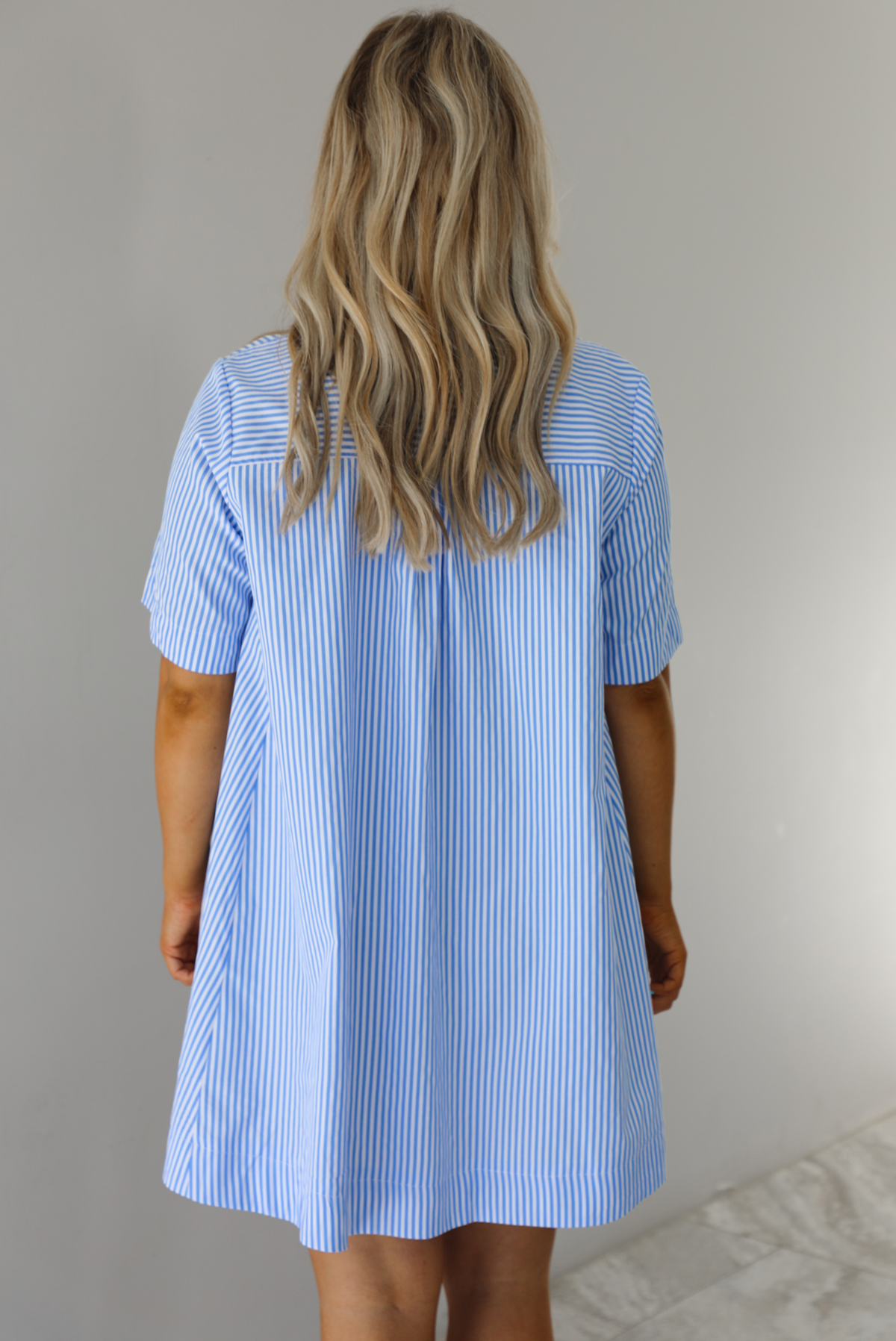 Woman wearing a blue and white striped dress that has a collared neckline, functional quarter button closures in the front, and a flowy skirt. The photo is taken indoors against a white wall and shows the back of the dress. 