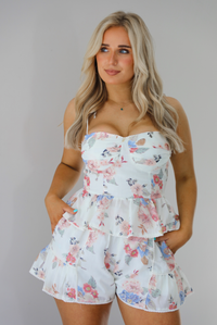 Woman wearing a white and multi-colored floral print patterned set. The photo is taken indoors against a white wall and shows a closeup of the top of the set, which has a corset-appearance with a fitted waist and a ruffled hem. 