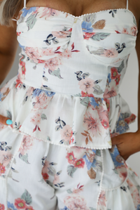 Woman wearing a white and multi-colored floral print patterned set. The photo is taken indoors against a white wall and shows a closeup of the top of the set, which has a corset-appearance with a fitted waist and a ruffled hem. 