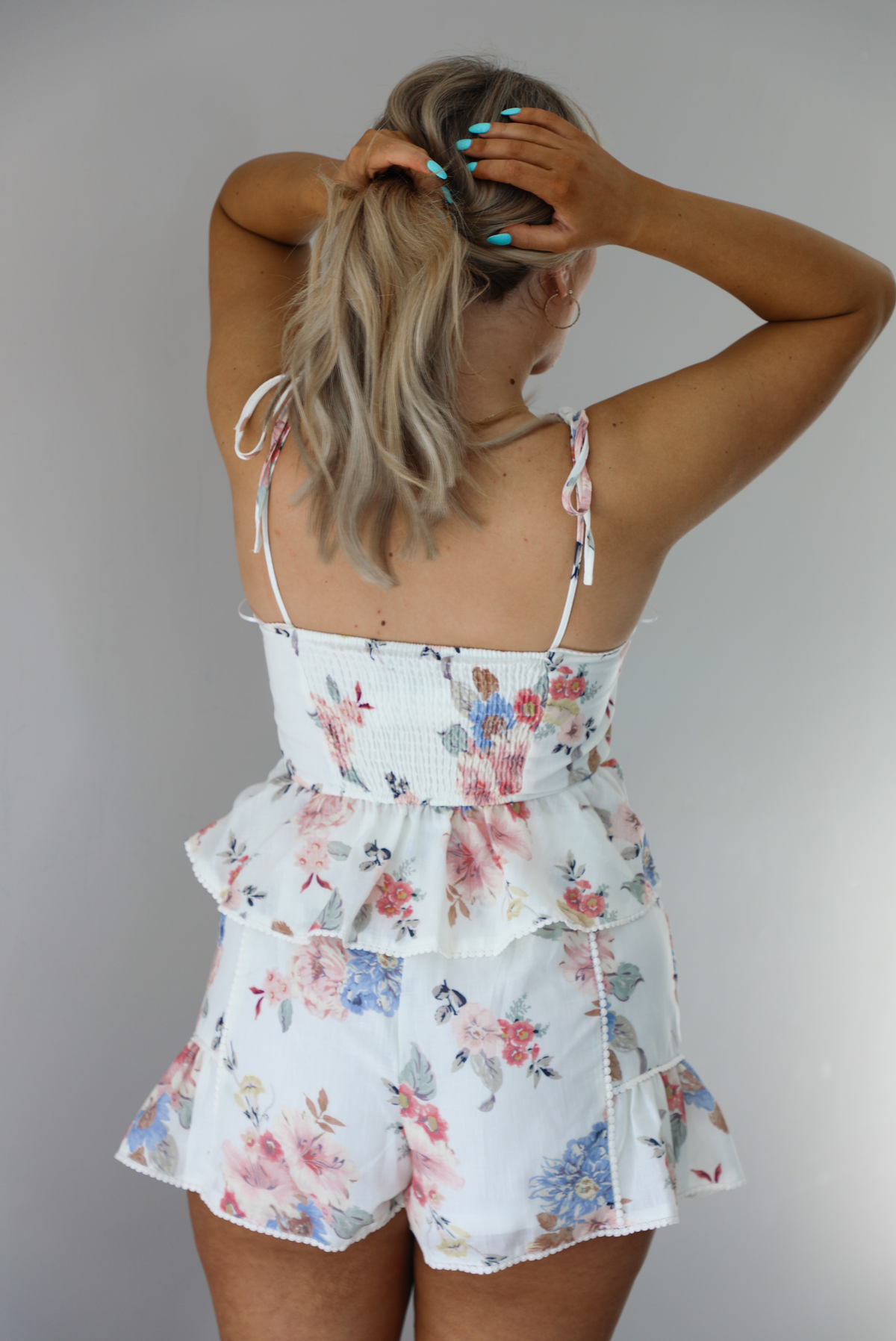 Woman wearing a white and multi-colored floral print patterned set. The photo is taken indoors against a white wall and shows a closeup of the back of the top of the set, which shows smocked detailing and adjustable straps. 