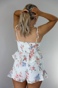 Woman wearing a white and multi-colored floral print patterned set. The photo is taken indoors against a white wall and shows a closeup of the back of the top of the set, which shows smocked detailing and adjustable straps. 
