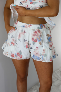 Woman wearing a white and multi-colored floral print patterned set. The photo is taken indoors against a white wall and shows a closeup of the shorts of the set. which are high waisted, have an elastic waistline, functional pockets on either side, and a ruffled hem. 