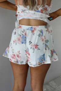 Woman wearing a white and multi-colored floral print patterned set. The photo is taken indoors against a white wall and shows a closeup of the back of the shorts of the set. which are high waisted, have an elastic waistline, functional pockets on either side, and a ruffled hem. 