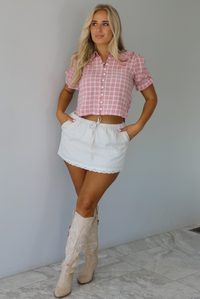 Woman wearing a pink and white plaid color throughout, has a collared neckline, functional button closures in the front, puffed sleeves, and a cropped cut. The top is worn with a low-rise grey skirt. The photo is taken indoors against a white wall with white marble flooring. 