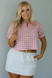 Woman wearing a pink and white plaid color throughout, has a collared neckline, functional button closures in the front, puffed sleeves, and a cropped cut. The top is worn with a low-rise grey skirt. The photo is taken indoors against a white wall.