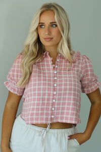 Woman wearing a pink and white plaid color throughout, has a collared neckline, functional button closures in the front, puffed sleeves, and a cropped cut. The top is worn with a low-rise grey skirt. The photo is taken indoors against a white wall.