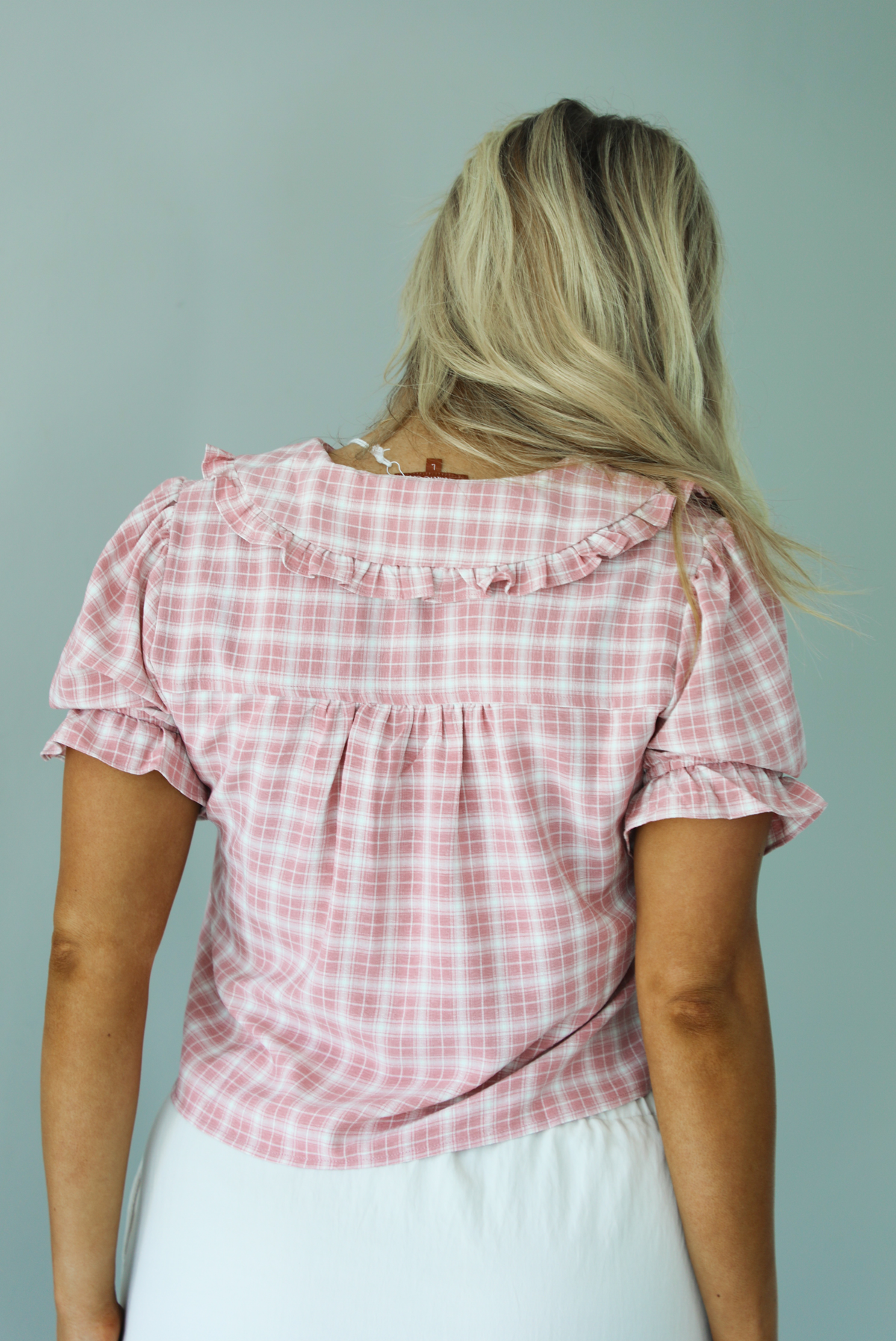 Woman wearing a pink and white plaid color throughout, has a collared neckline, functional button closures in the front, puffed sleeves, and a cropped cut. The top is worn with a low-rise grey skirt. The photo is taken indoors against a white wall and shows the back of the top. 