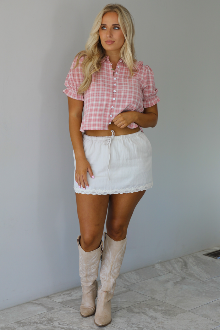 Woman wearing a pink and white plaid color throughout, has a collared neckline, functional button closures in the front, puffed sleeves, and a cropped cut. The top is worn with a low-rise grey skirt. The photo is taken indoors against a white wall with white marble flooring. 