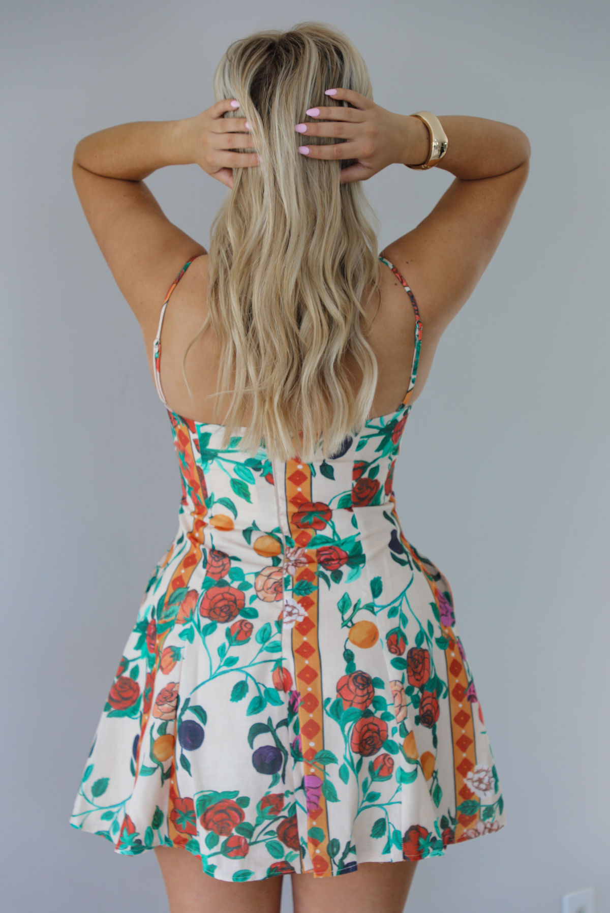 Woman wearing a high neck floral mini dress with thin straps, functional pockets on either side, and a flowy skirt. The photo is taken indoors against a white wall and shows the back of the dress, which has adjustable straps and a zipper closure. 
