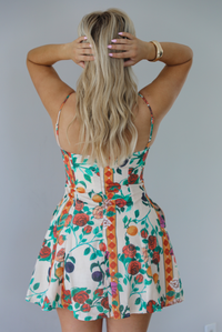 Woman wearing a high neck floral mini dress with thin straps, functional pockets on either side, and a flowy skirt. The photo is taken indoors against a white wall and shows the back of the dress, which has adjustable straps and a zipper closure. 