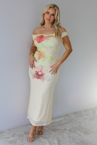 Woman wearing a cream colored, off the shoulder maxi dress that has a multi-colored floral print pattern throughout with a cut out on the right side. The photo is taken indoors against a white wall with white marble flooring. 
