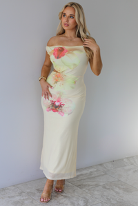 Woman wearing a cream colored, off the shoulder maxi dress that has a multi-colored floral print pattern throughout with a cut out on the right side. The photo is taken indoors against a white wall with white marble flooring. 