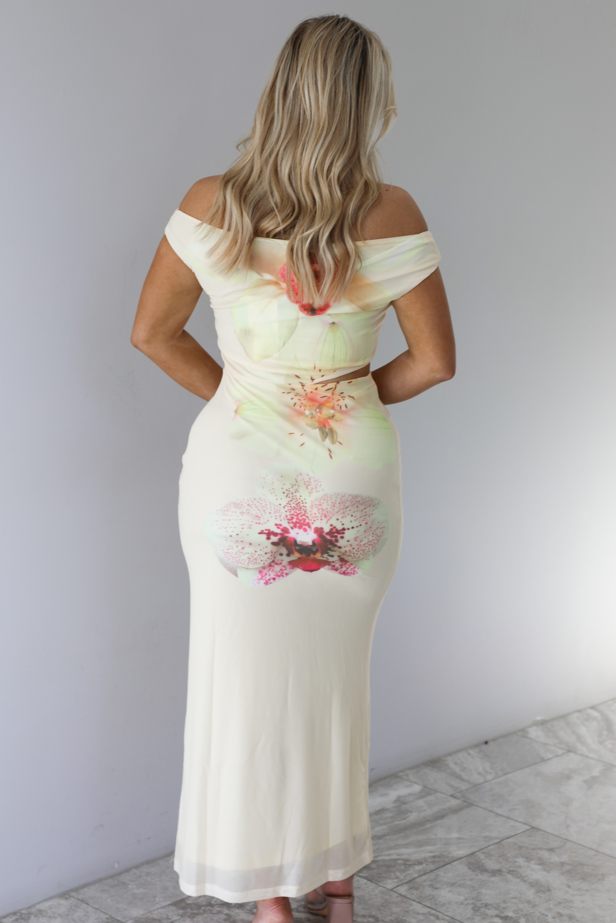 Woman wearing a cream colored, off the shoulder maxi dress that has a multi-colored floral print pattern throughout with a cut out on the right side. The photo is taken indoors against a white wall with white marble flooring and shows the back of the dress. 