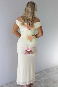 Woman wearing a cream colored, off the shoulder maxi dress that has a multi-colored floral print pattern throughout with a cut out on the right side. The photo is taken indoors against a white wall with white marble flooring and shows the back of the dress. 