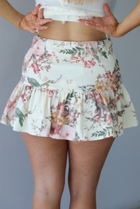 Woman wearing a cream and multi-colored floral skirt that has a high waistline, and a shorter length. The photo is taken indoors against a white wall and shows the back of the skirt. 
