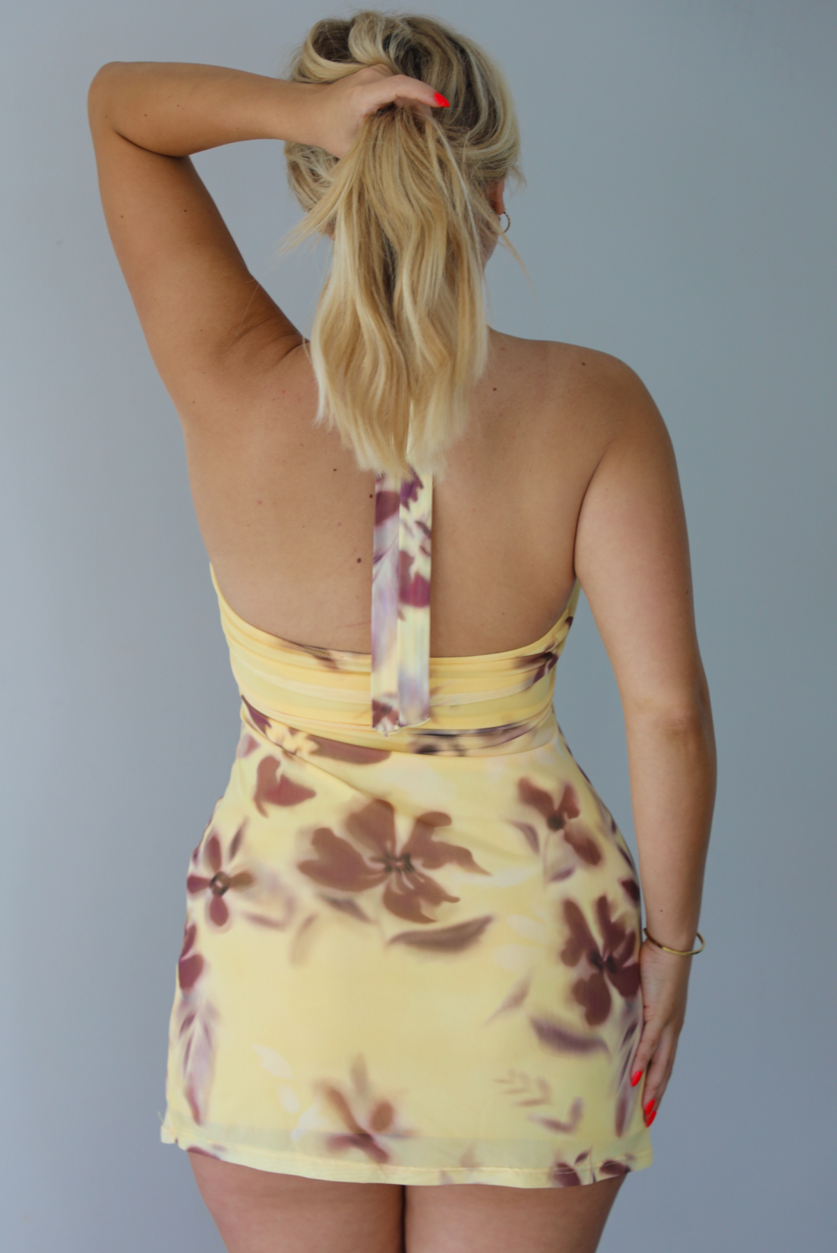Woman wearing a yellow dress with a brown floral print pattern that has a halter tie neckline and a mini length. The photo shows the back of the dress and is taken indoors against a white wall. 