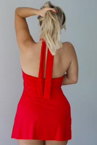 Woman wearing a red mini dress with straps that connect at the center of the top of the dress and tie around the neck. The photo is taken indoors against a white wall. The photo shows the back of the dress. 