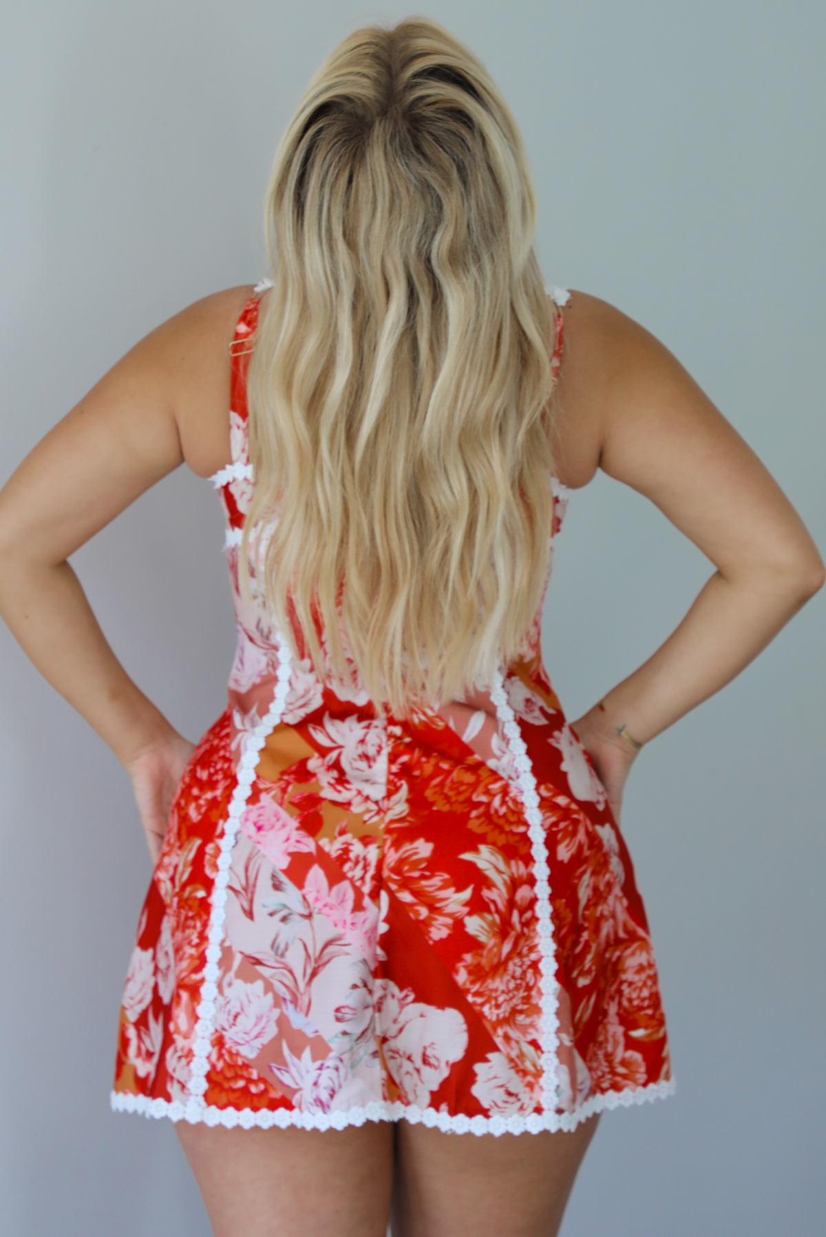 Woman wearing a red, orange, and white floral mini dress with white trim detailing indoors against a white wall. The photo shows a closeup of the back of the dress, which has a zipper closure and functional adjustable straps. 