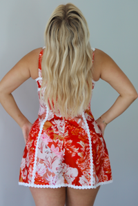 Woman wearing a red, orange, and white floral mini dress with white trim detailing indoors against a white wall. The photo shows a closeup of the back of the dress, which has a zipper closure and functional adjustable straps. 