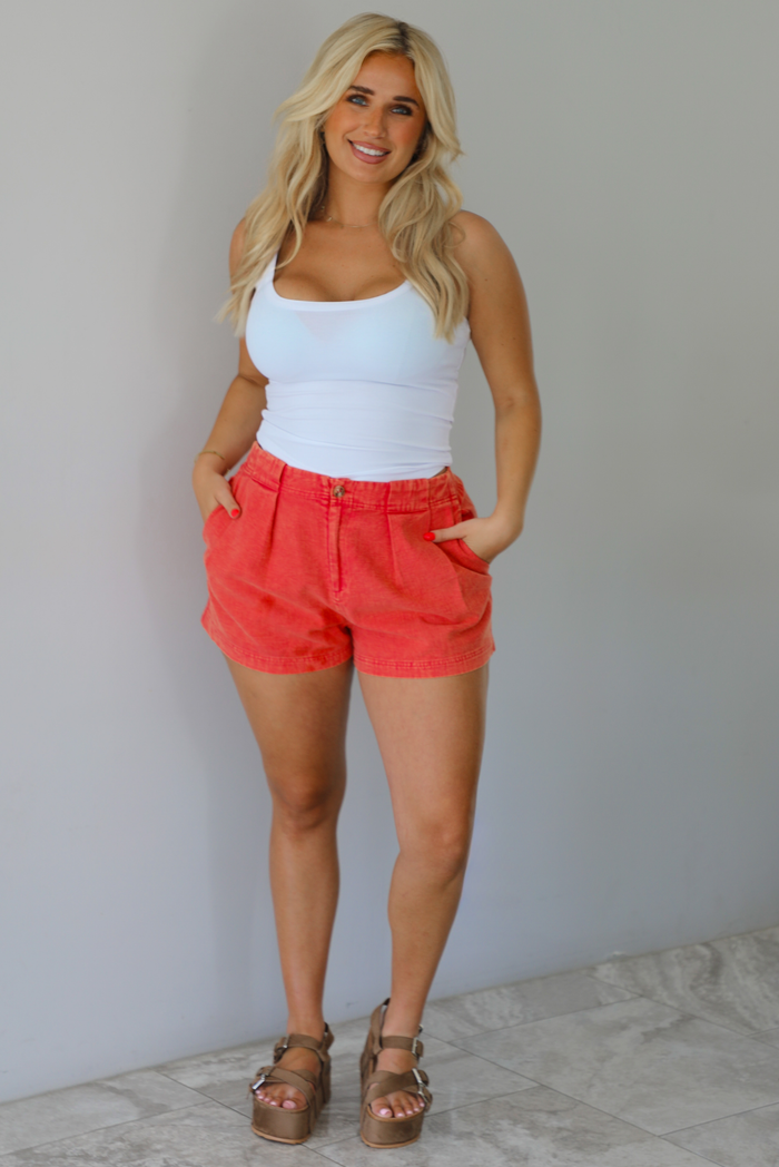 Woman wearing a white tank top and red shorts indoors against a white wall with white marble flooring. 