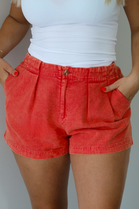 Woman wearing a white tank top and red shorts indoors against a white wall. The photo shows a closeup of the shorts with the models hands in the pockets on either side, and the pleated details in the front shown. 
