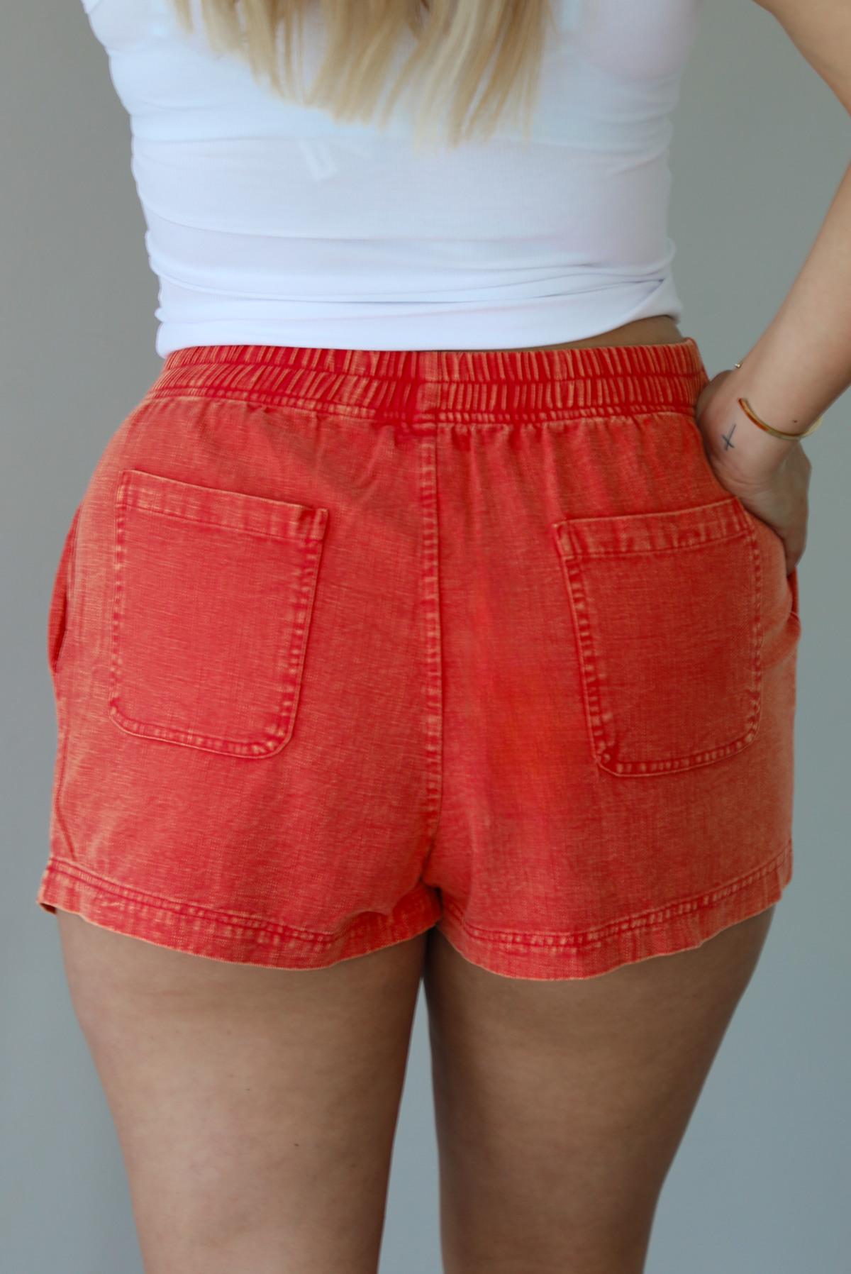 Woman wearing a white tank top and red shorts indoors against a white wall. The photo shows the back of the shorts, which features an elastic waistline and functional back pockets. 
