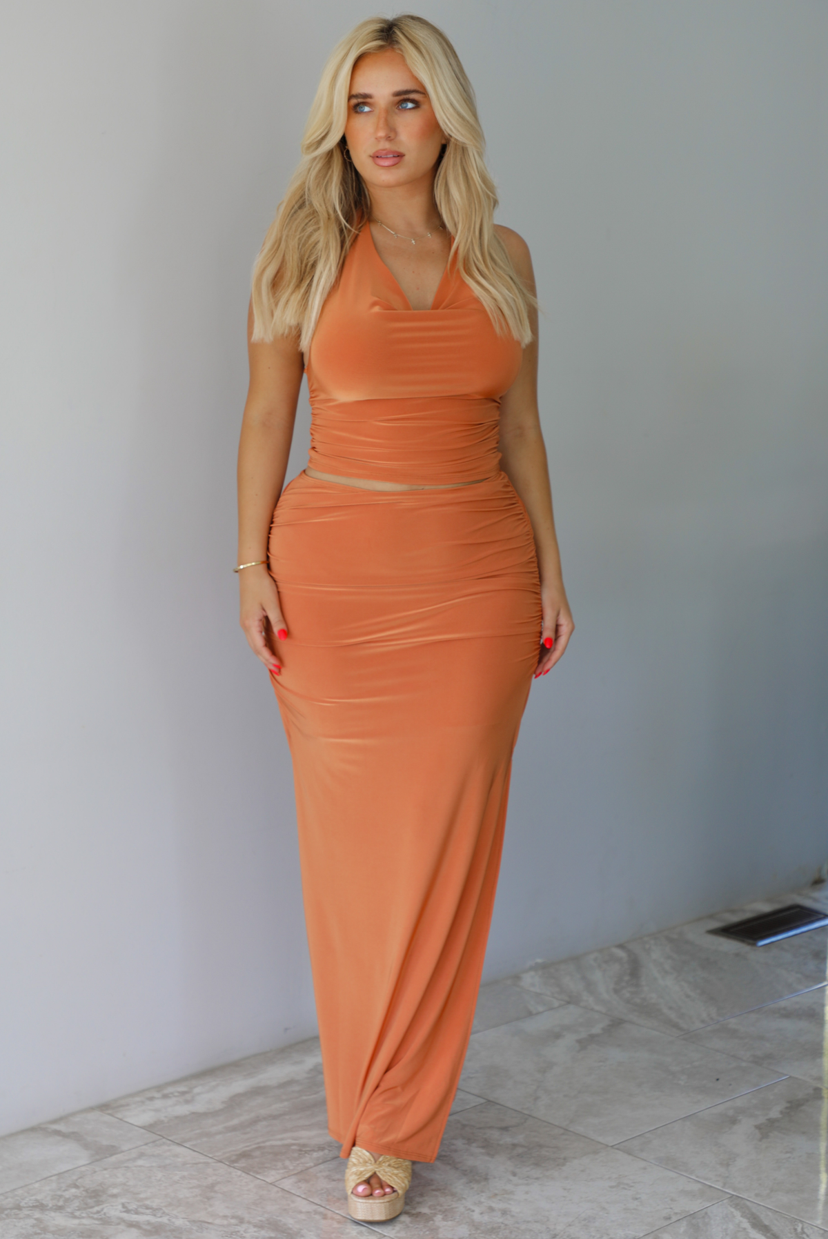 Woman wearing an orange halter neck and maxi-length skirt indoors against a white wall with white marble flooring. 