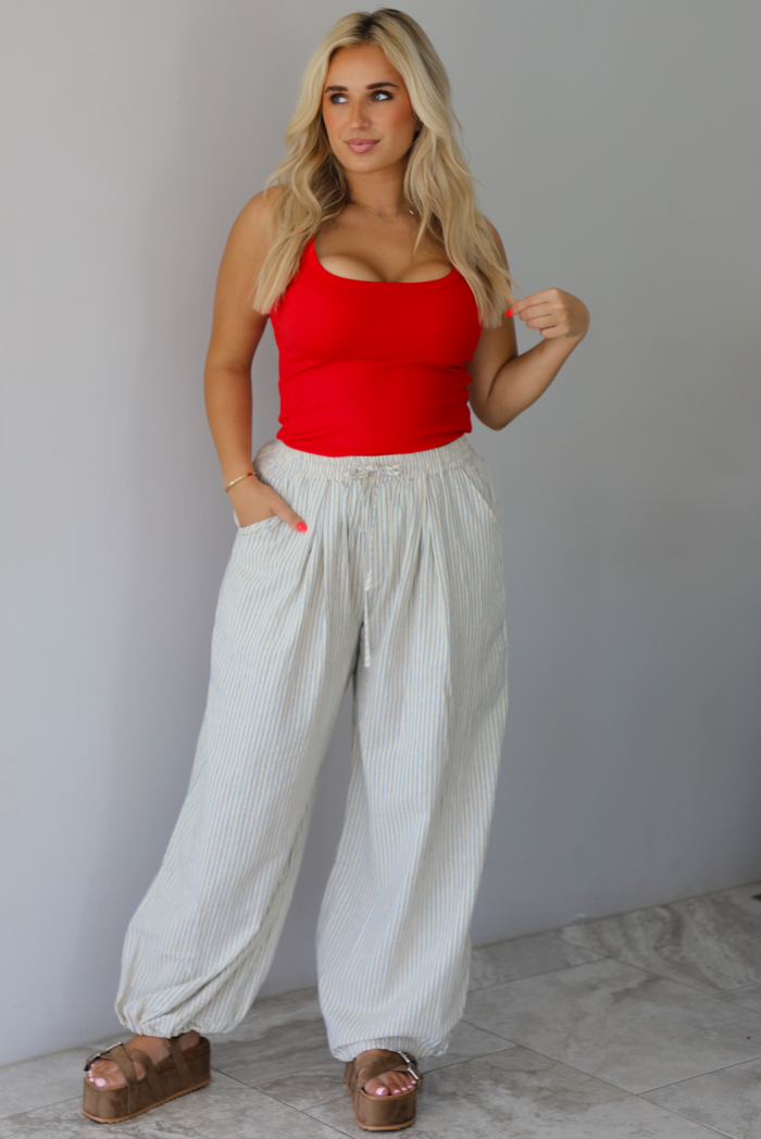 Woman wearing a red tank tucked info ecru lounge pants that have a blue striped design, high waistline, and functional pockets on either side indoors against a white wall with white marble flooring. 