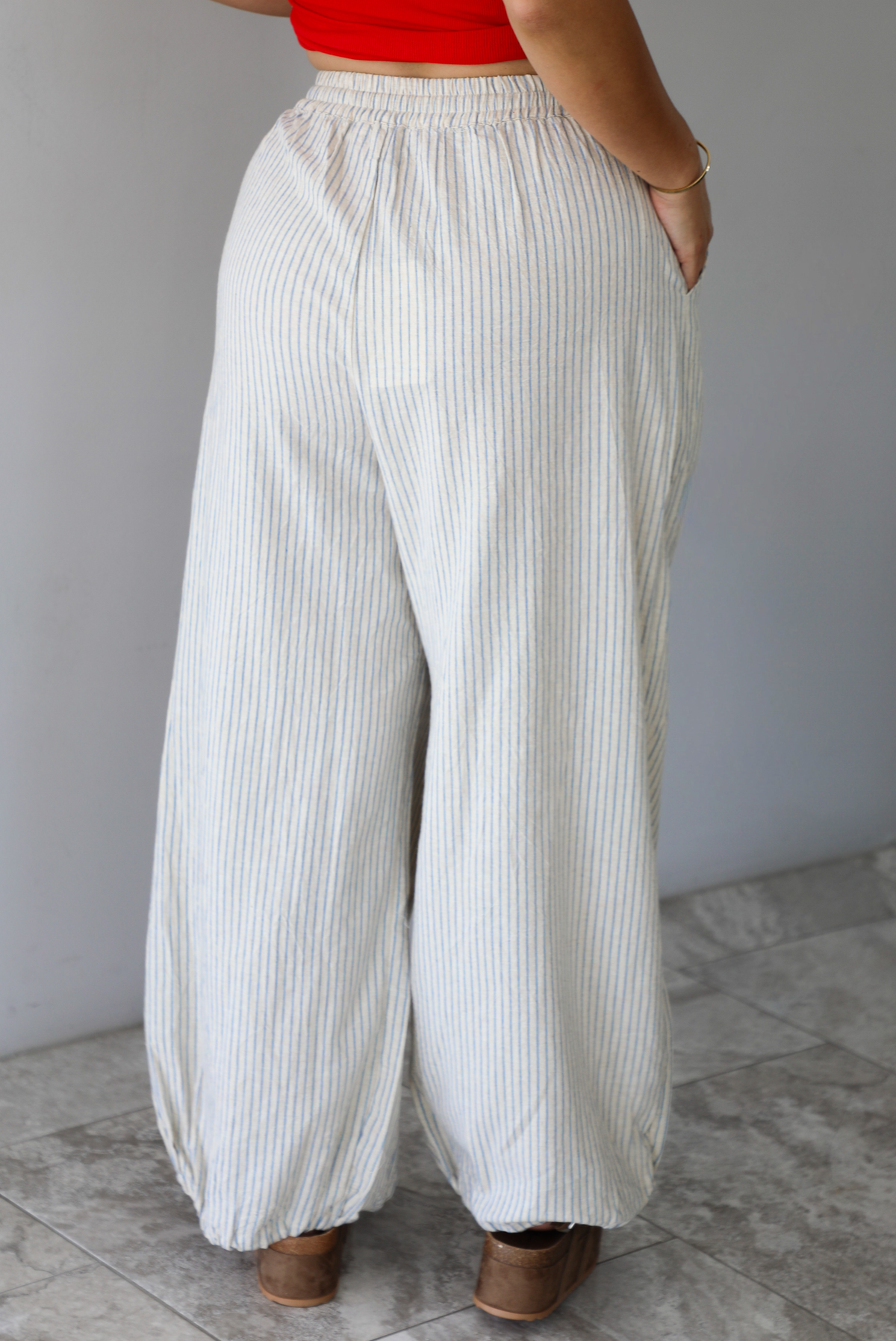 Woman wearing lounge pants that are an ecru color with a blue striped pattern, are high waisted with an elastic and drawstring waistline, and have a slouched design until they cinch at the ankles indoors against a white wall with white marble flooring. The photo shows the back of the pants. 