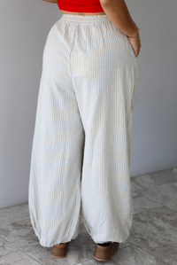 Woman wearing lounge pants that are an ecru color with a blue striped pattern, are high waisted with an elastic and drawstring waistline, and have a slouched design until they cinch at the ankles indoors against a white wall with white marble flooring. The photo shows the back of the pants. 